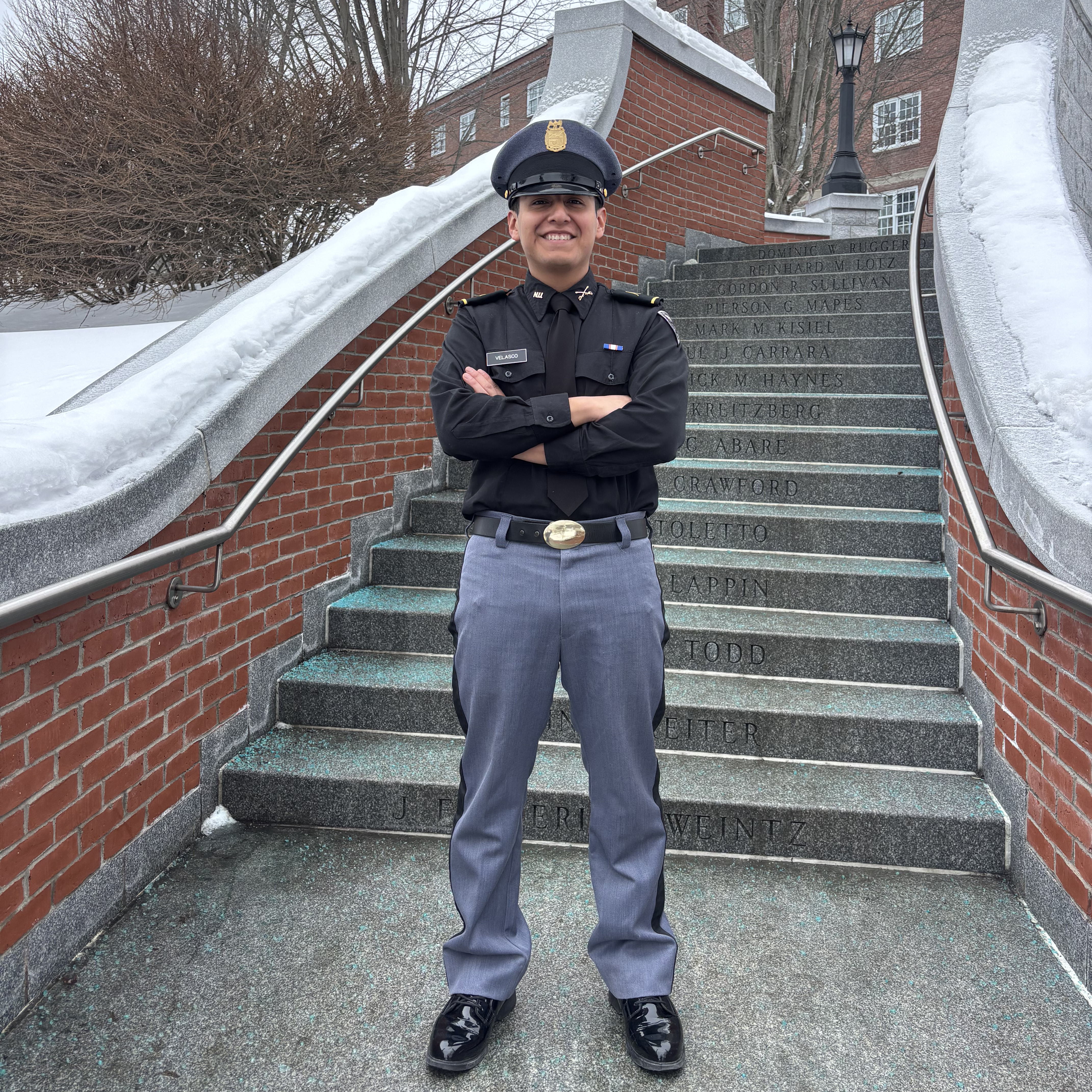Person in a dark uniform and cap stands with arms crossed on outdoor stone steps bordered by brick walls and snow.