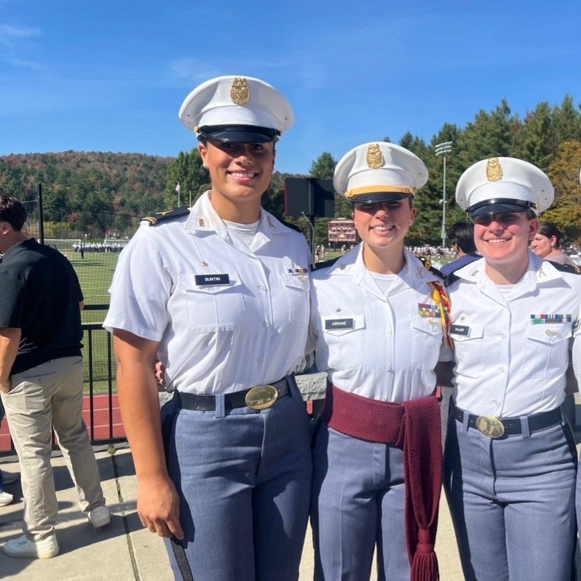 Four cadets in white dress uniforms stand together beside a football field on a sunny day.