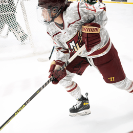 A hockey player in a military-themed camouflage uniform competing against opponent in green and yellow stripes, with teammates visible in the background.