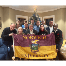 Norwich University flag held by twelve people, all standing in front of a Christmas tree