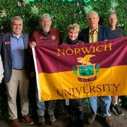 A group of individuals stand before a greenery covered wall. Four individuals in the middle hold up a maroon and gold Norwich University flag.