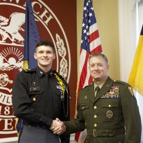 BG William F. McCollough, VSM, Commandant and Vice President for Student Affairs, Norwich Class of 1991 and C/1SG Matthew S. Ober '26 shaking hands in front of the Norwich University logo