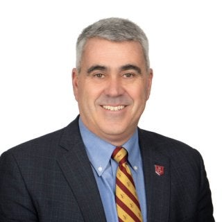 A man with short gray hair wears a plaid jacket, light blue shirt, maroon & golds striped tie and an N logo pin, standing against a white background.