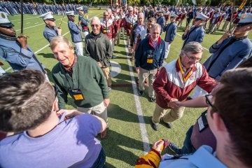 Two men, on left in green henley shirt, on right, in a red & white jacket, approaching other individuals to shake hands. 