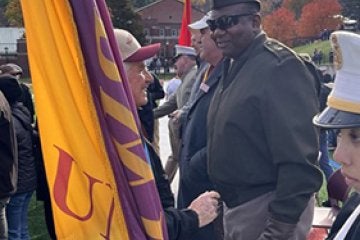 A person holding a yellow and purple flag speaks with a person in a military-style uniform at an outdoor gathering.