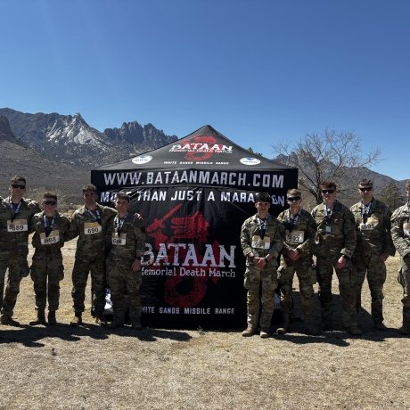 The Norwich University cadets who participated in the event stand shoulder-to-shoulder in front of a branded tent in the desert with a mountain range in the background.
