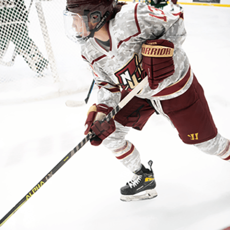 A hockey player in a military-themed camouflage uniform competing against opponent in green and yellow stripes, with teammates visible in the background.