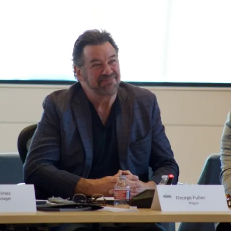 Person seated at a conference table, smiling, between name plates labeled Paul Grimes, City Manager, and George Fuller, Mayor.