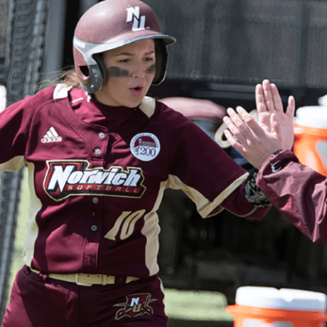 Norwich softball player in maroon uniform and batting helmet receiving high-five from coach near dugout, wearing number 10 jersey.