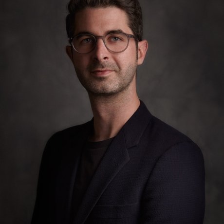 Portrait of an individual with glasses wearing a dark blazer, set against a textured grey background.