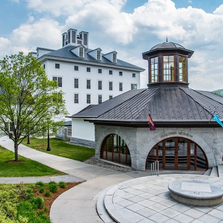 Exterior view of the Sullivan Museum and History Center at Norwich University, featuring a unique curved building with a tall cupola in front and a large multi-story building in the background, set against a backdrop of lush greenery and clear skies.