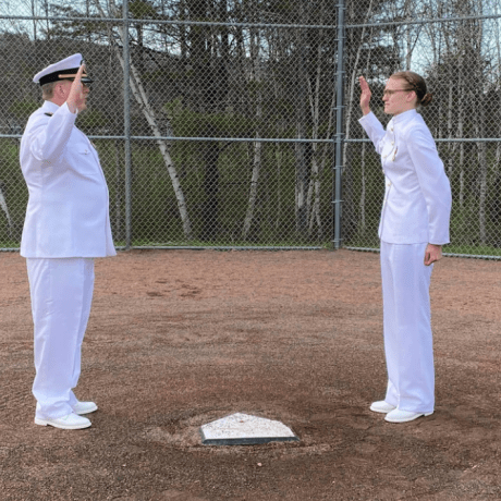 Caitlin Dailey (right) stands next to home plate while her father (left, directly across home plate) swears her in. They are both in full white Navy outfits.