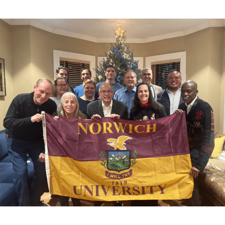 Norwich University flag held by twelve people, all standing in front of a Christmas tree