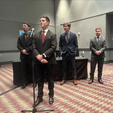 Four people in suits stand in a room with patterned carpet, one speaking at a microphone while others stand behind.