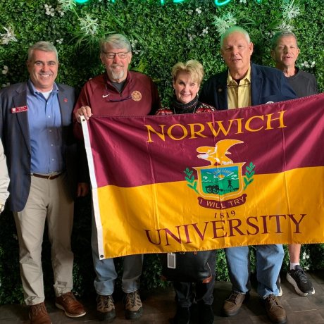 A group of individuals stand before a greenery covered wall. Four individuals in the middle hold up a maroon and gold Norwich University flag.