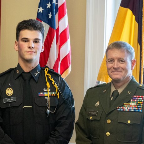 Two people in military dress uniforms stand indoors in front of U.S. and yellow-and-brown flags.