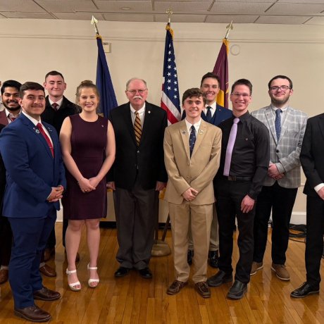 A group of people in formal clothing stand in rows on a wooden floor with three flags displayed behind them.