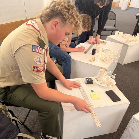 Person in a scout uniform measures a sheet with a ruler at a white table covered with cups, tape, notes, and small materials.