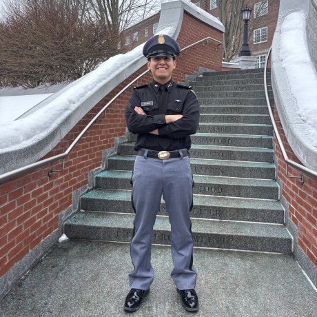 Person in a dark uniform and cap stands with arms crossed on outdoor stone steps bordered by brick walls and snow.
