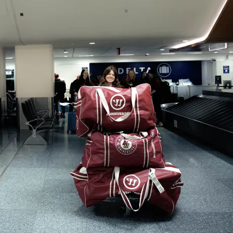 Student pushes cart stacked with Norwich University duffel bags through an airport baggage claim area.