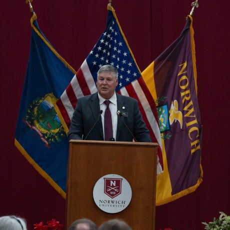 Person speaks at a Norwich University podium with Vermont, U.S., and Norwich flags behind.