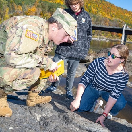 Cadet and students examine rocks beside a river with autumn trees and a bridge in the background.