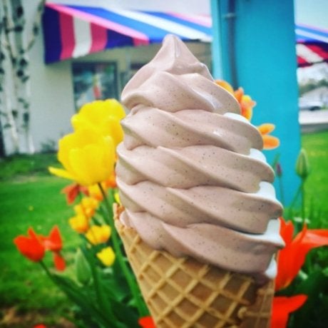 The top swirl of a creemee / soft-serve ice cream and cone, with flowers, building with colorful awning in background.