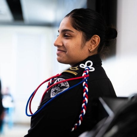 Cadet in dress uniform with red, white, and blue shoulder cords looks toward the front of a room during a