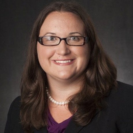 A person wearing glasses and a pearl necklace poses against a dark background.