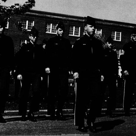 Cadets in uniform march in formation outside a brick building.