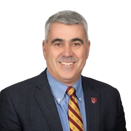 A man with short gray hair wears a plaid jacket, light blue shirt, maroon & golds striped tie and an N logo pin, standing against a white background.
