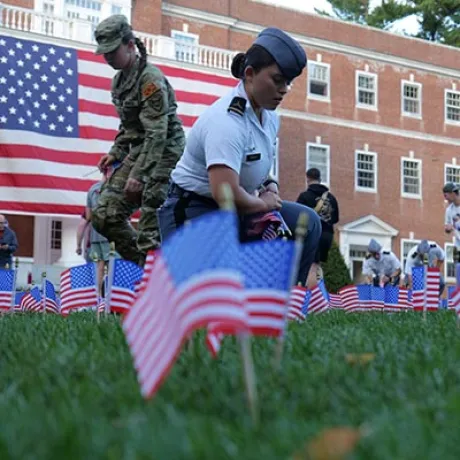 Cadets place small U.S. flags in the grass in front of a large U.S. flag and campus building.