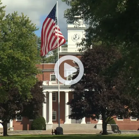 American flag flies in front of a brick building with white columns and a clock tower.
