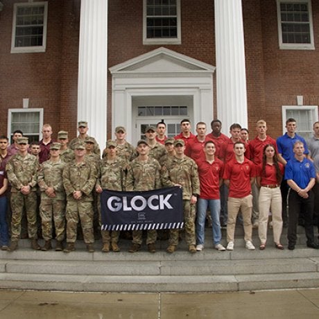 Cadets in camouflage uniforms and students in colored polo shirts pose with a GLOCK banner on steps outside a brick building.