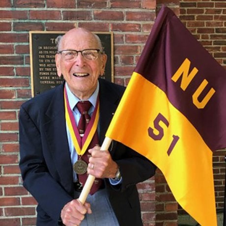 Portrait of R. Damon holding a maroon and gold Norwich University Class of 1951 flag while wearing a medal.