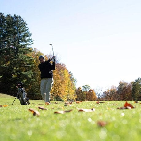 A golfer swings a club on a grassy course with autumn trees in the background.