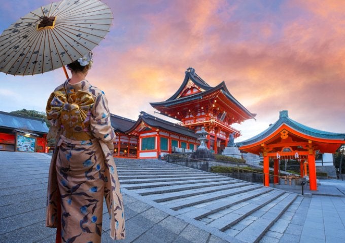 A woman in japanese dress, holding an umbrella, looks upon several pagoda buildings.