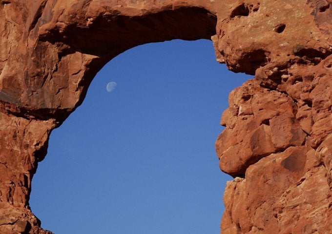 Arch geological feature in US Southwest frames sky and glimpse of partial full moon.