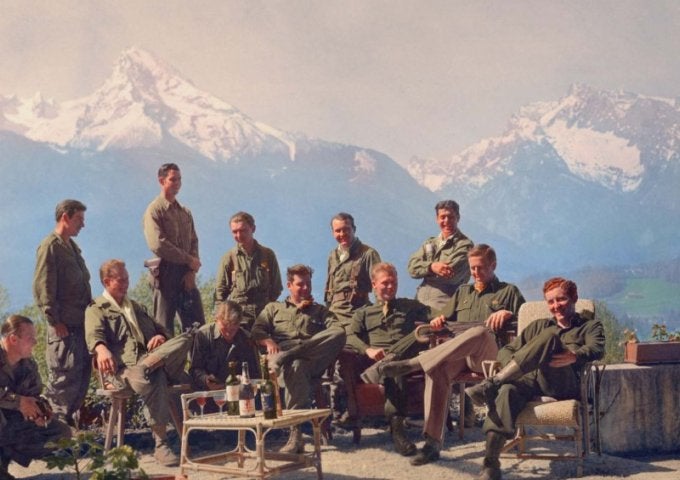 Ten service members in uniform sit and stand together on a terrace with snow-covered mountains in the background.