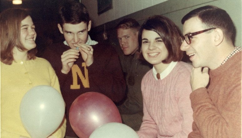 Four people indoors holding balloons, one person inflating a balloon.