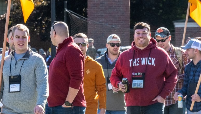 Seven individuals in casual attire walk forward, several holding up maroon and gold flags (guidons).