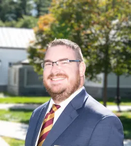 Person in a suit and tie poses against a plain light background wearing a Norwich pin.