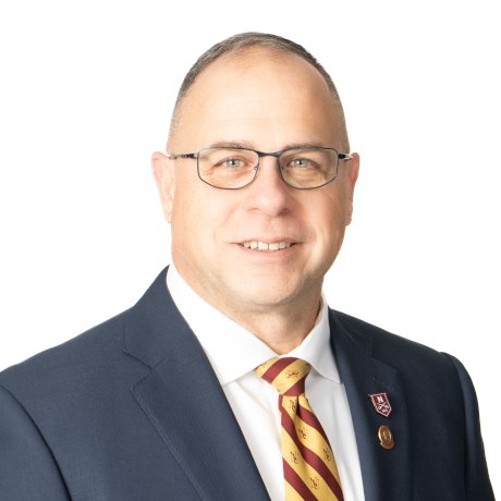 Person wearing glasses, in a dark gray suit, maroon and gold striped tie, and two lapel pins.