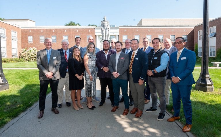 A group of individuals in business attire stand in a courtyard with brick single-story building in the background.