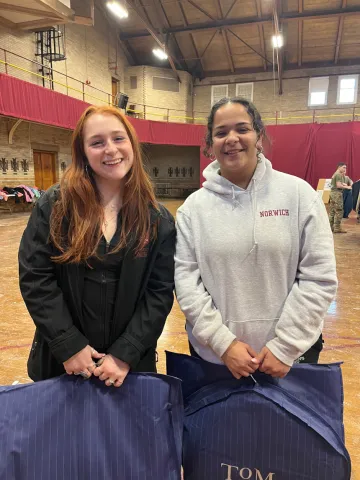 Two women standing beside each other, one at left wearing green jacket and shirt, the other at right wearing a light gray hoodie sweatshirt with small "Norwich" lettering top right. They are holding formal wear donated to them through Dressing for Success, held Feb. 9-10 at Norwich University.