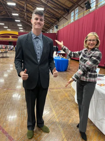 A young man in dark suit stands next to a women; he's giving a thumb's up while she motions as if to present him to the photographer. They are both wearing formal wear donated to him through Dressing for Success, held Feb. 9-10 at Norwich University.
