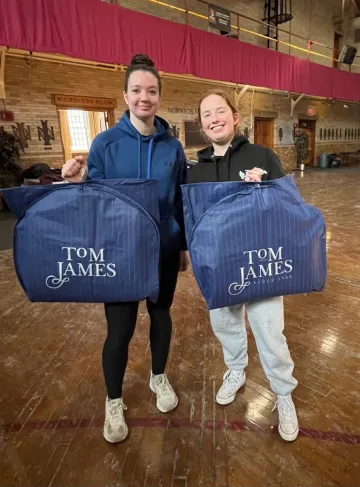 Two women standing beside each other. both wearing hooded sweatshirts. They hold blue pinstriped garment bags that say "Tom James" containing formal wear donated to them through Dressing for Success, held Feb. 9-10 at Norwich University.