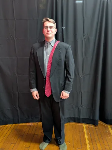 A young man in gray suit and slacks poses, with his arms at his sides and a bright red tie draped over his left shoulder. He is donning formal wear donated to him through Dressing for Success, held Feb. 9-10 at Norwich University.