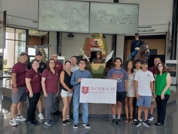 A small group of young adults stand in middle surrounded by older adults. Two in middle hold up a modestly sized white banner reading "Norwich University".