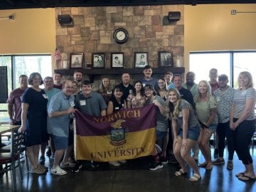 A group of young adults stand in foreground with a line of older adults beside them. Two individuals hold a Norwich University flag at width ends, and a fireplace and mantel are in the background.